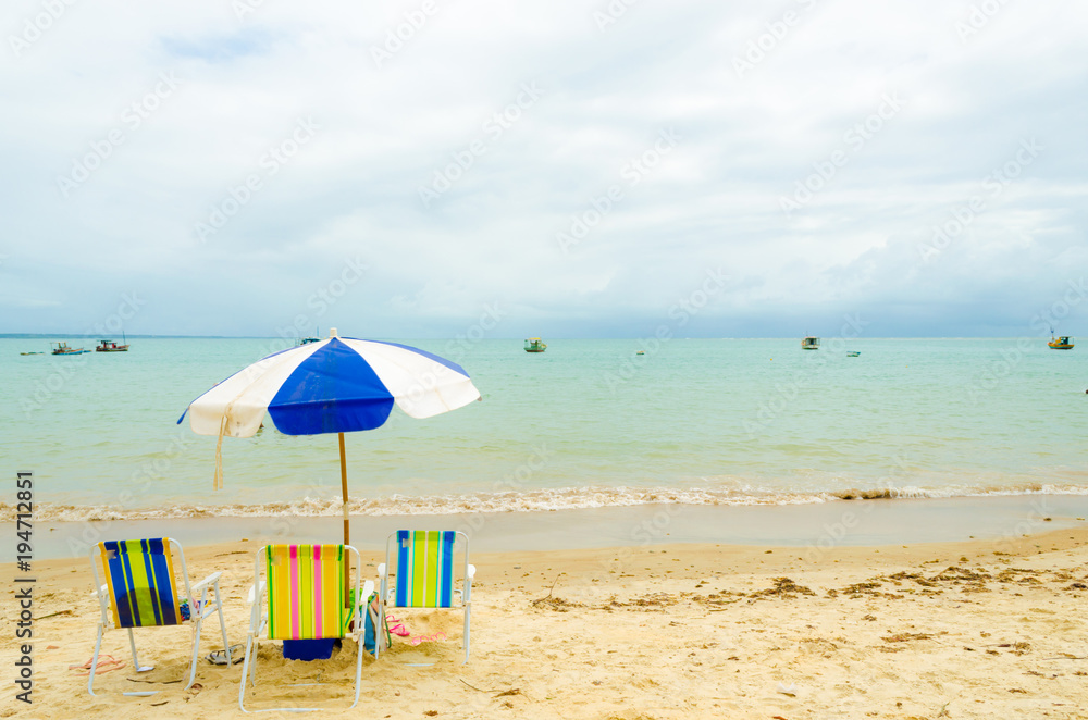 Isolated beach with three colorful beach chairs and a blue and white umbrella. A small wave breaks in the sand and some boats are in the sea. Cloudy sky.