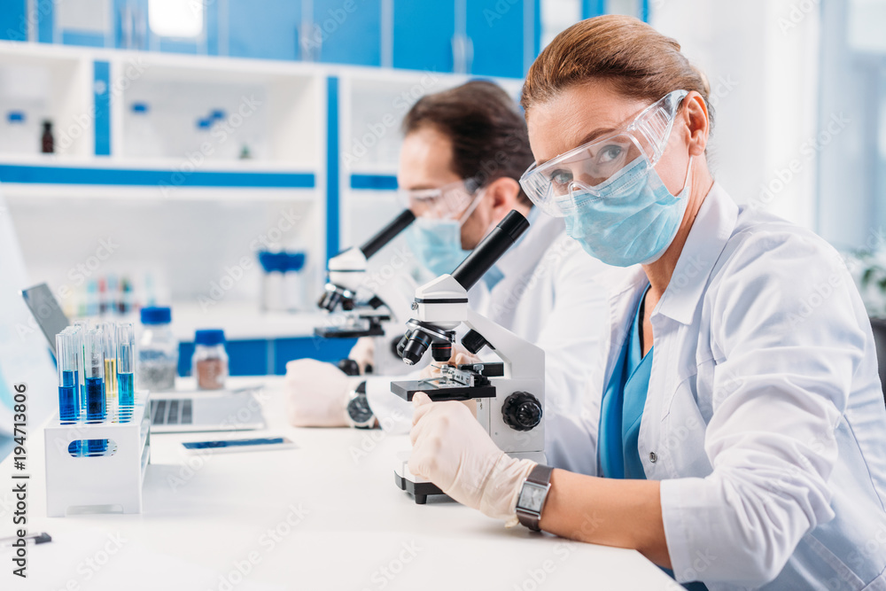 selective focus of scientists in medical masks and goggles looking through microscopes on regents in lab selective focus of scientists in medical masks and goggles looking through microscopes on regents in lab