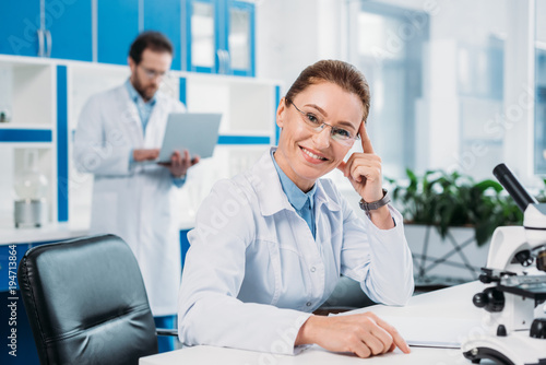 selective focus of smiling female scientist in eyeglasses at workplace with colleague behind in lab