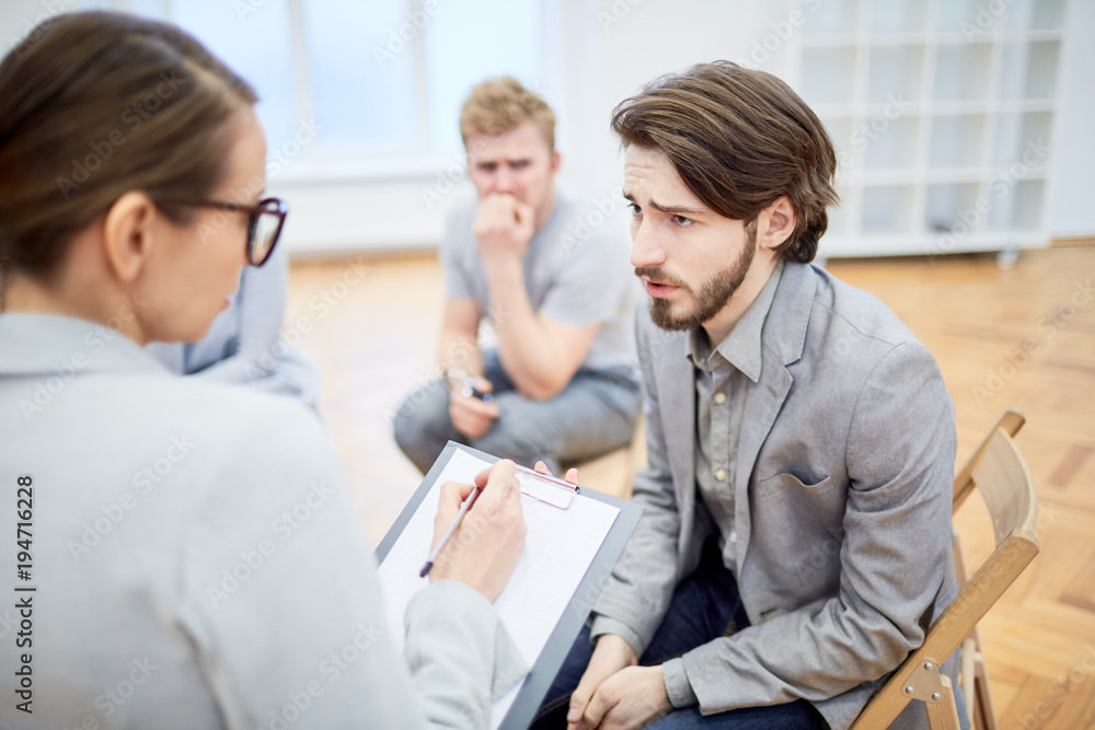 Troubled young businessman explaining his problem to counselor during group session Troubled young businessman explaining his problem to counselor during group session