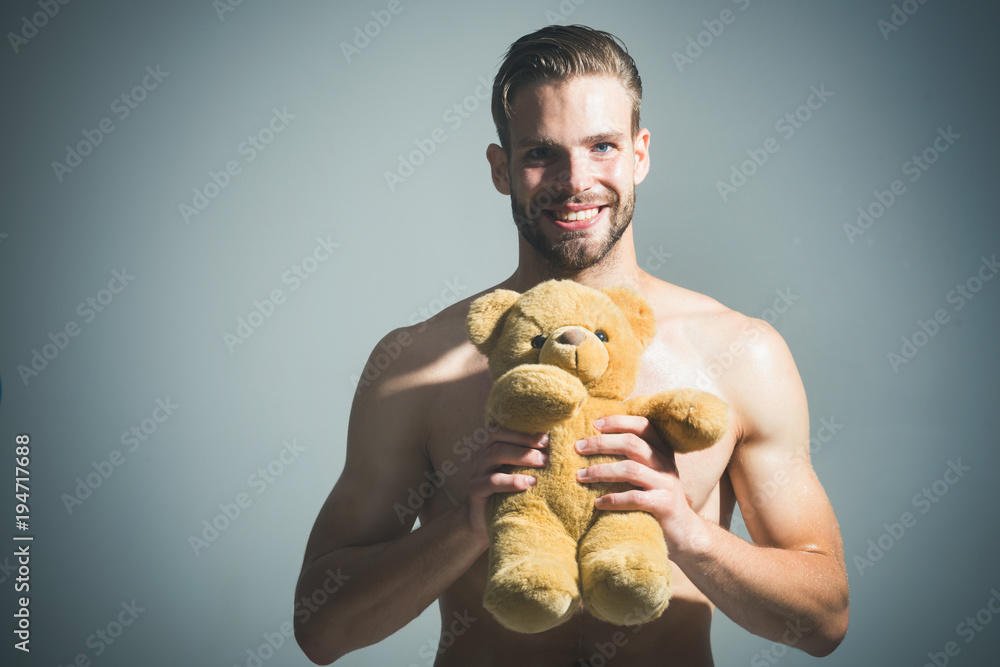 Young sexy attractive guy with toy bear. Handsome man with his teddy bear. Smiling bearded man holding teddy bear. Cute young adult, happy man is holds teddy bear in hands, smiles, on gray background.