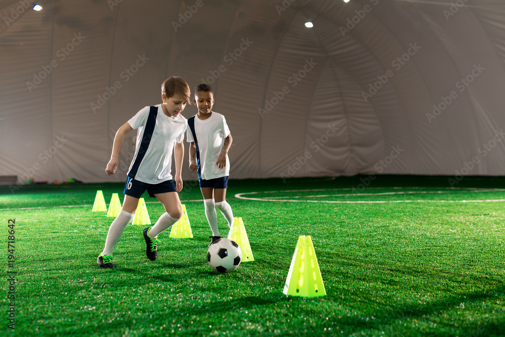 Fototapeta premium Two little football players running after ball while exercising on pitch with cones