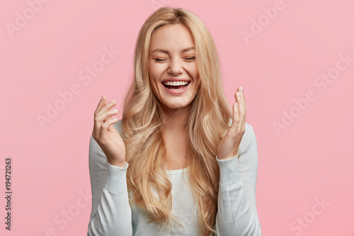Fototapeta Isolated shot of joyful blonde young cute woman laughs joyfully as hears funny anecdote from friend, has long light hair, poses against pink studio wall