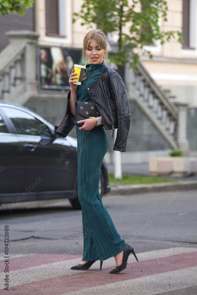 Fototapeta premium Beautiful rich luxury looking blonde caucasian business woman in casual green outfit standing on a european streets, holding coffee and purse. Daylight, outdoors.