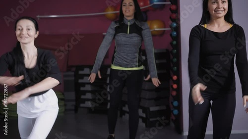 three brunette women are doing bodily exercises in a gymnastic hall, looking forward and repeating for coach