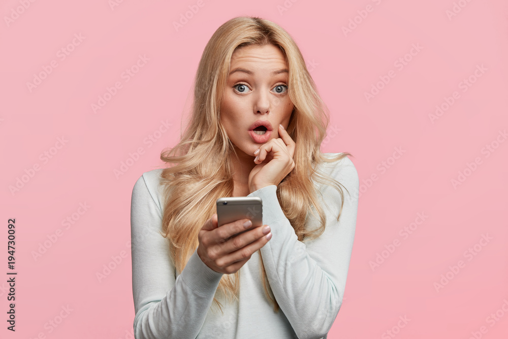Beautiful female with blonde hair feels surprised as checks her email box on cell phone, wonders big discounts in web store, isolated over pink background. Emotions and unexpectedness concept Beautiful female with blonde hair feels surprised as checks her email box on cell phone, wonders big discounts in web store, isolated over pink background. Emotions and unexpectedness concept