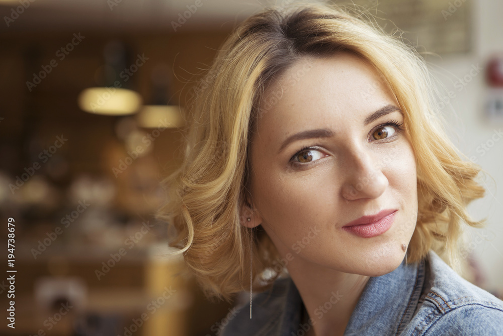 Fototapeta premium Blonde (with dark roots) caucasian woman in casual summer outfit at the cafe. Grey dress and jeans jacket. Woman got natural day makeup and curly hairstyle. Indoors