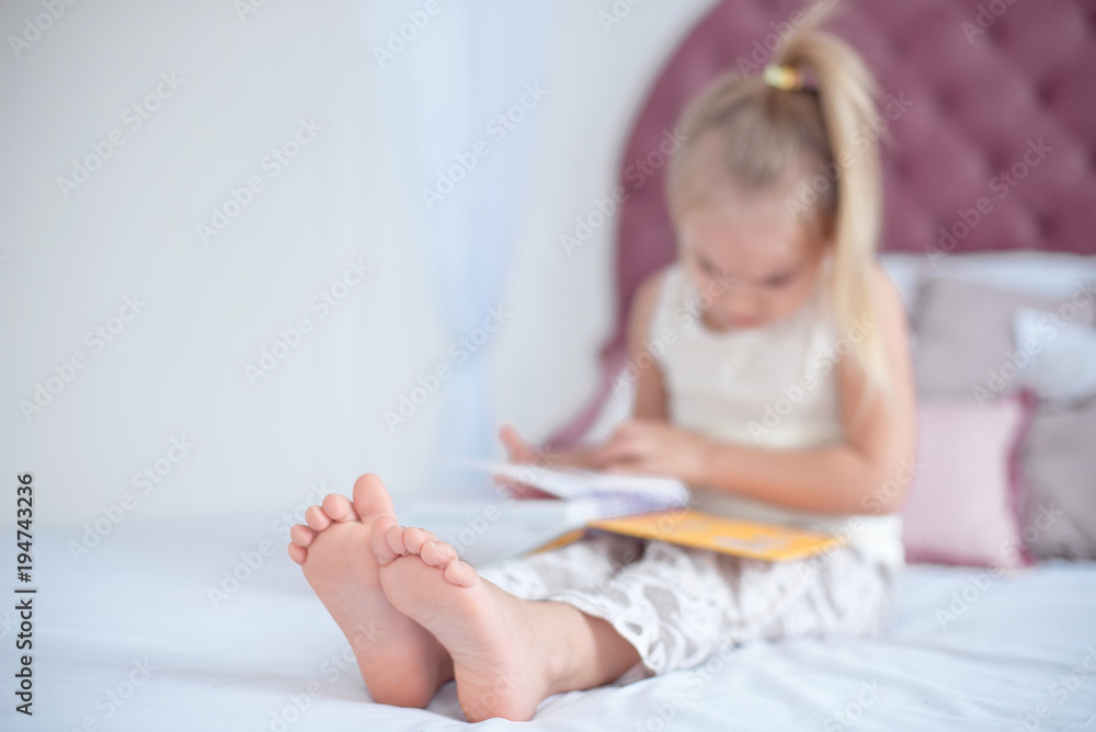 Little blonde girl sitting on the bed reading a book with focus on the feet  indoors Stock Photo | Adobe Stock
