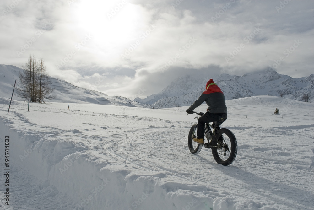 Fototapeta premium man use electric bicycle, e-bike, ebike, pedal on snow covered road, downhill mountain, specific bike with wide wheels to go on snow, called fatbike, winter, cold, alps, Simplon Pass, Switzerland