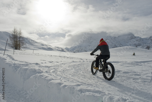 man use electric bicycle, e-bike, ebike, pedal on snow covered road, downhill mountain, specific bike with wide wheels to go on snow, called fatbike, winter, cold, alps, Simplon Pass, Switzerland