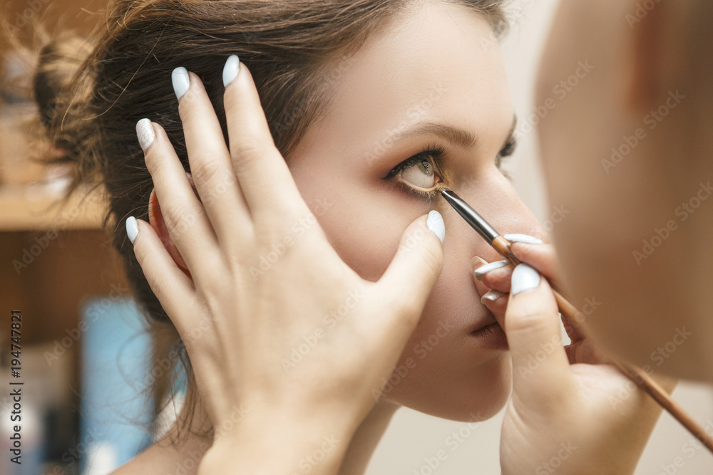 Obraz premium Make up artist woman applying make up for a brunette model for a photosession. Painting her eyes with an eye shadows using a brush. Nude lips, clear skin