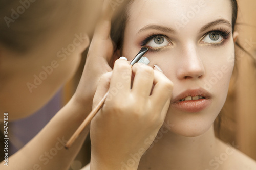 Make up artist woman applying make up for a brunette model for a photosession. Painting her eyes with an eye shadows using a brush. Nude lips, clear skin