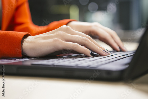 Business woman sitting in her modern office in luxury red jacket. Neutral interior with dim led lighting. Woman working on a laptop, close up of her hands typing on a keyboard