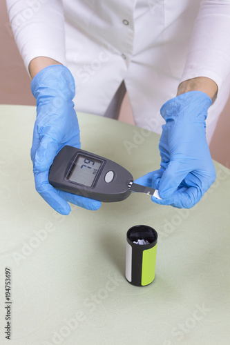 Ręka w rękawiczce jednorazowej wyciąga pasek do mierzenia poziomu cukru we krwi glukometrem z pudełka. Glukometr leży na stole.