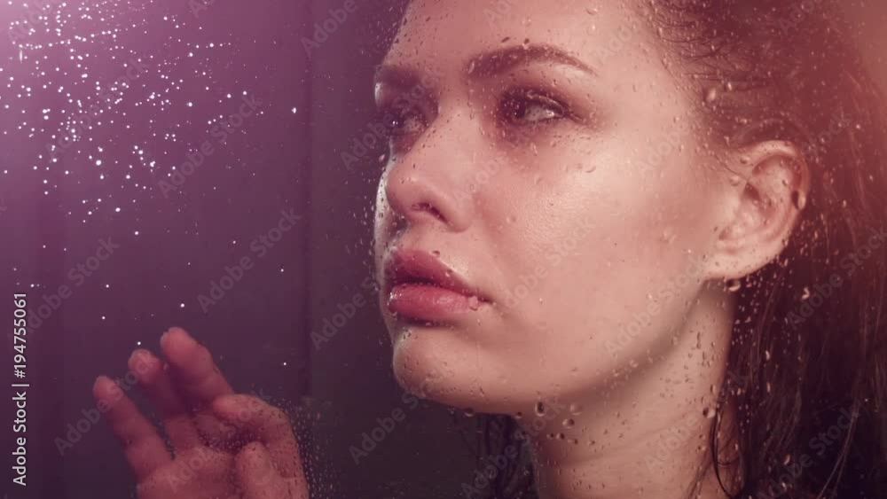 Beautiful girl with wet hair behind the glass under shower. Naked girl takes a dush. Drops water flow down glass. Thoughtful expression. Means for skin care. Girl dreams looking at rain behind glass. Beautiful girl with wet hair behind the glass under shower. Naked girl takes a dush. Drops water flow down glass. Thoughtful expression. Means for skin care. Girl dreams looking at rain behind glass.