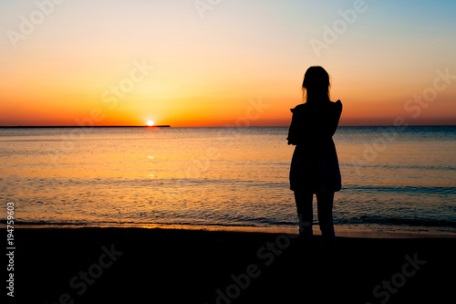 Woman in summer dress standing on a sandy beach and looking to the sun