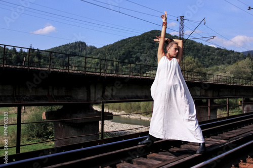 Young beautiful girl in a long beautiful white dress. The brunette poses to the photographer against the background of a beautiful landscape. A girl in a white outfit in good sunny weather.