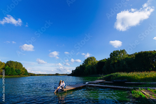 A woman with a wreath on her head and her husband are sitting on the pier of a beautiful lake