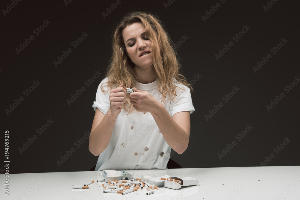 Waist up portrait of angry blonde woman sitting at table with pile of cigarettes and squashing several of them with hands. Isolated on background Waist up portrait of angry blonde woman sitting at table with pile of cigarettes and squashing several of them with hands. Isolated on background