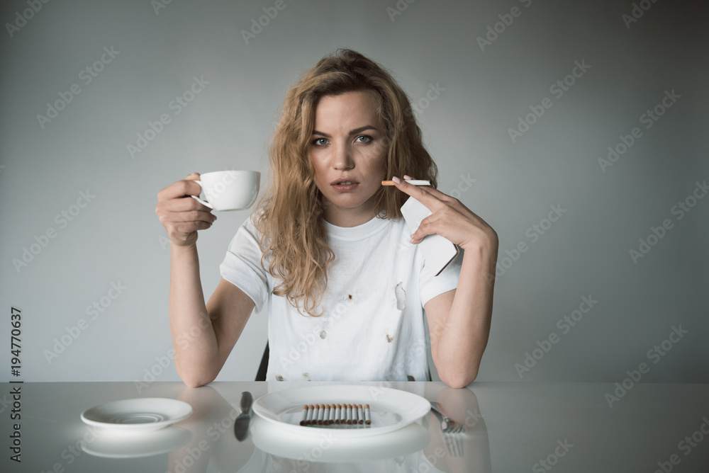 Portrait of serious girl holding mug in one hand and cellphone with ciggy  in another. She is sitting and looking at camera. Isolated on background  Stock Photo | Adobe Stock