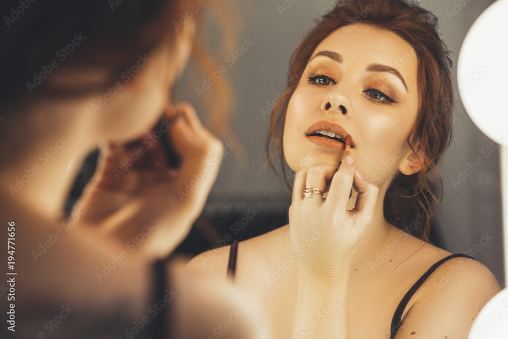 Brunette woman applying make up (paint her lips) for a evening date in front of a mirror. Focus on her reflection Brunette woman applying make up (paint her lips) for a evening date in front of a mirror. Focus on her reflection