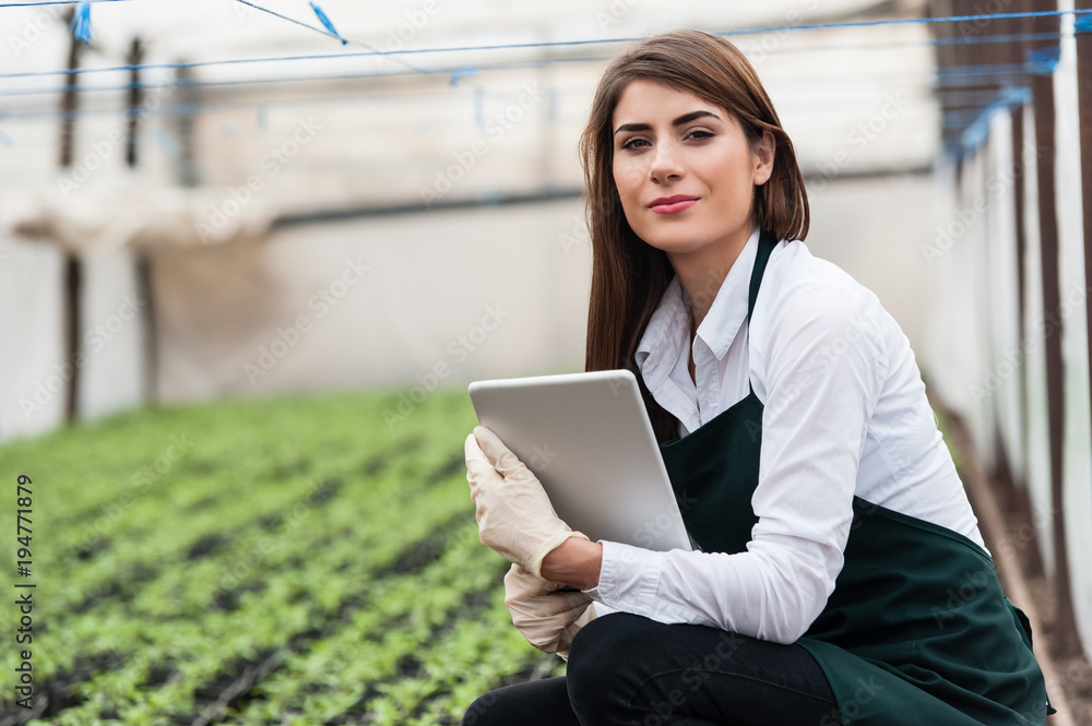 Beautiful young biotechnology woman engineer with clipboard and pen, writing and examining plants for disease in greenhouse with apron and gloves. Greenhouse produce. Food production.