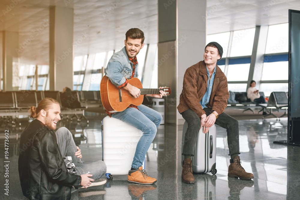 Naklejka premium Cheerful male playing musical instrument near friends with luggages in airport. Expectation concept