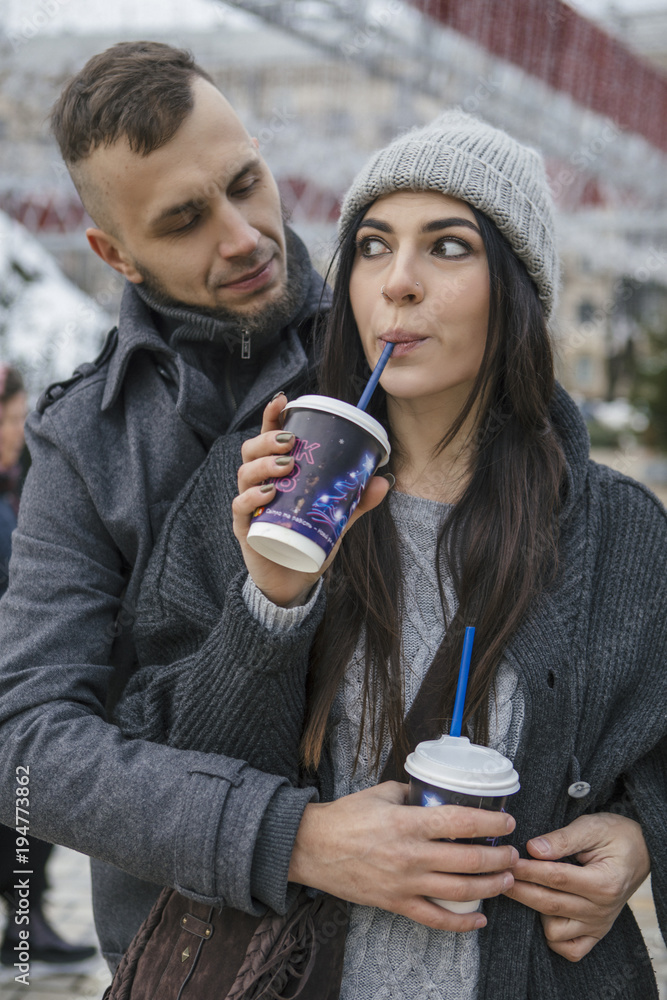 Fototapeta premium Gorgeous beautiful caucasian woman with a man on a walk in european city at winter. Couple wear casual grey outfit and hugging each other. Cold weather, christmas lights on a background