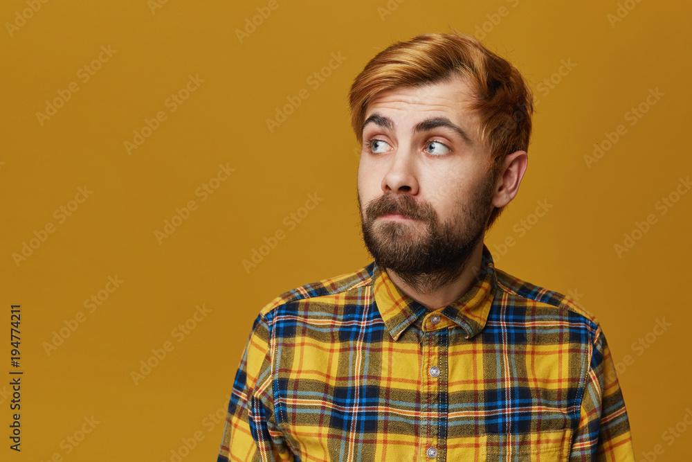 Young hipster curving his lip wears plaid yellow shirt looking aside. Serious expression. Isolated over yellow background with copyspace Young hipster curving his lip wears plaid yellow shirt looking aside. Serious expression. Isolated over yellow background with copyspace