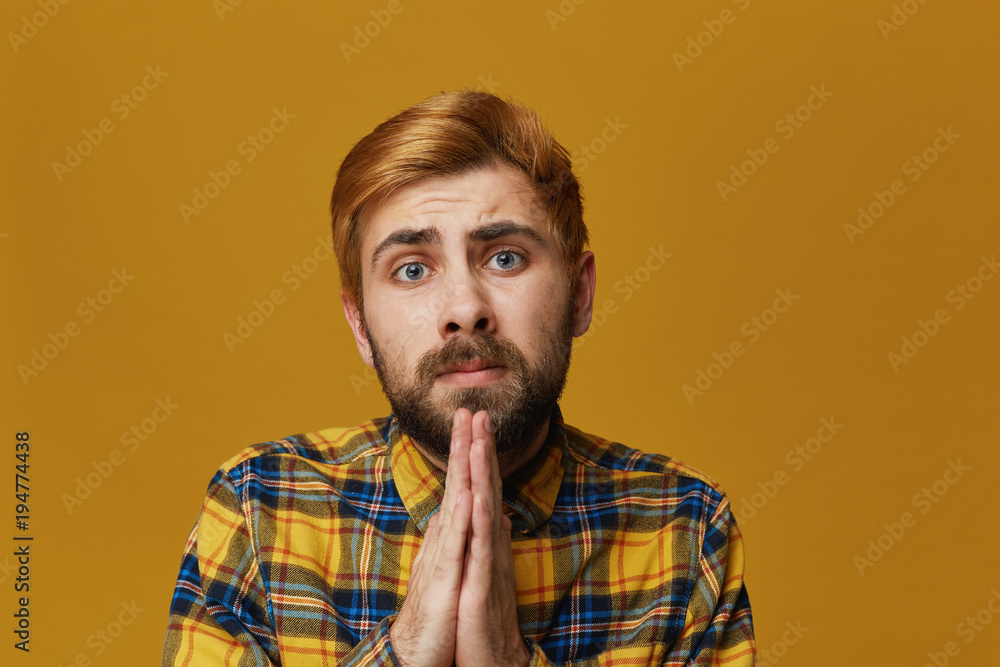 Obraz premium Portrait of serious young bearded male, keep his palms together, prays for good luck on exams. Isolated over yellow background.