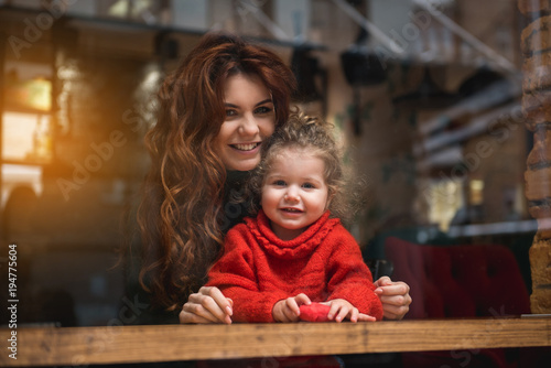 Portrait of happy mother embracing her little daughter while sitting at table. They are looking forward over the window and smiling