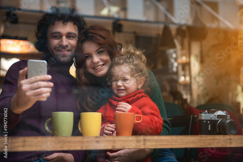 Lets remember this moment. Portrait of joyful young family making selfie on smartphone and smiling. They are sitting at table in cafeteria and cuddling