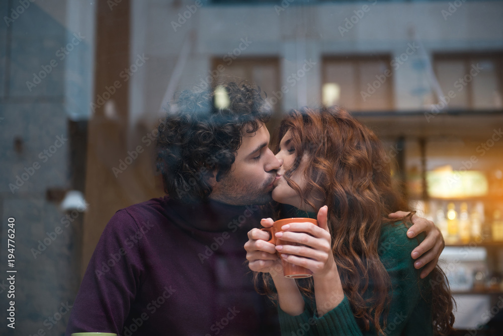 Fototapeta premium Happy loving couple kissing with gentleness. They are embracing while girl is holding mug of hot coffee. View from the glass window