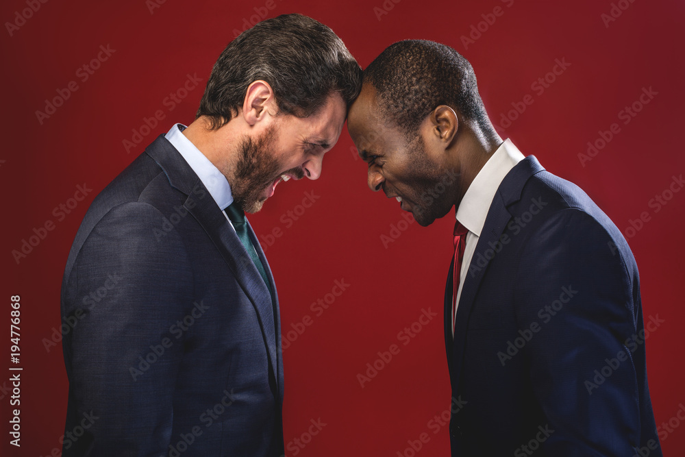Side view profile of angry young colleagues are fighting each other with foreheads together, staring with hostile expressions. Isolated on red background Side view profile of angry young colleagues are fighting each other with foreheads together, staring with hostile expressions. Isolated on red background