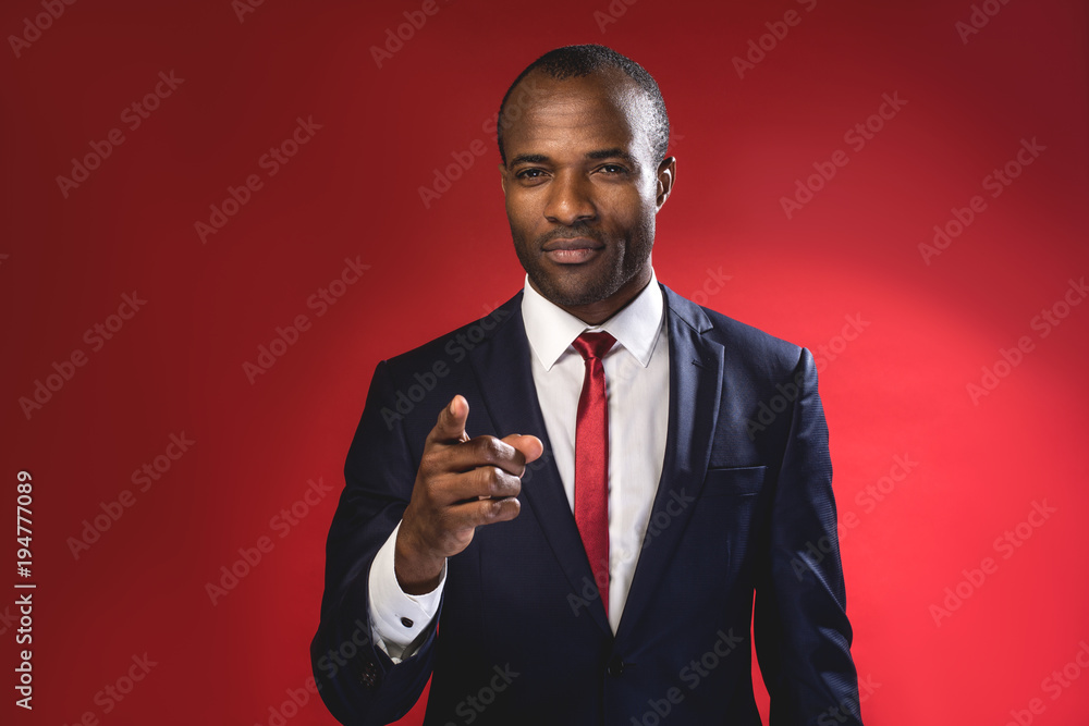 Hey you. Waist-up portrait of serious young african manager is pointing you while standing. Isolated on red background Hey you. Waist-up portrait of serious young african manager is pointing you while standing. Isolated on red background