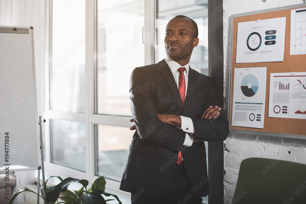 Confident expert. Portrait of confident young african manager is looking aside pensively. He is standing in office with crossed arms Confident expert. Portrait of confident young african manager is looking aside pensively. He is standing in office with crossed arms