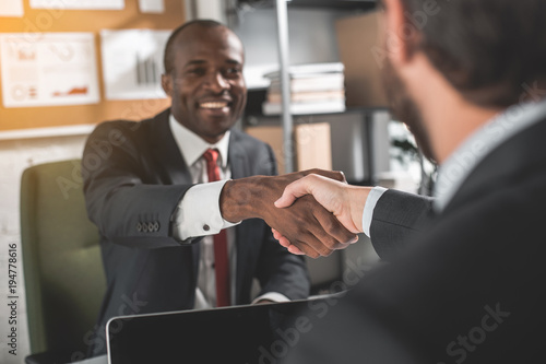 Good work. Portrait of cheerful african businessman is sitting at table with his partner. They are having strong handshake as finalize an agreement. Selective focus