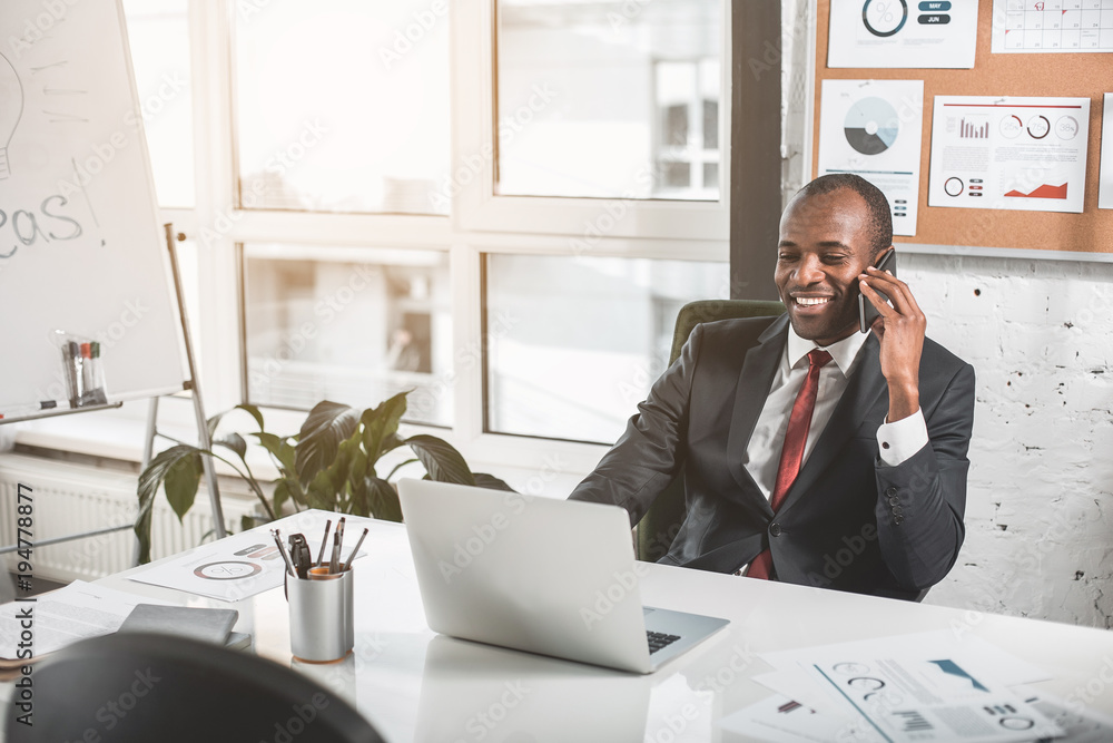 Good news. Cheerful young african businessman is talking with his business partner on mobile phone with smile. He is sitting at table in office and working on laptop. Copy space in the left side