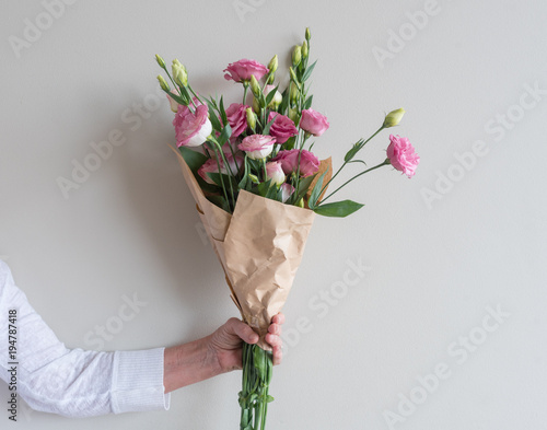 Woman's hand holding bouquet of pink lisianthus flowers wrapped in in brown paper against neutral wall background
