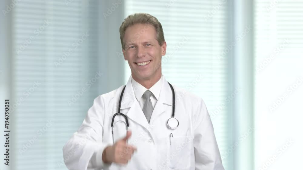 Joyful cardiologist showing thumb up. Positive middle-aged doctor putting on stethoscope and gesturing thumb up, blurred background. Joyful cardiologist showing thumb up. Positive middle-aged doctor putting on stethoscope and gesturing thumb up, blurred background.