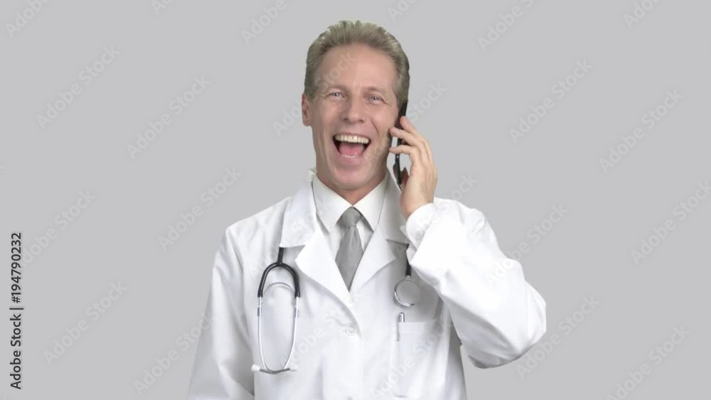 Excited male doctor talking on phone. Cheerful cardiologist talking on mobile phone and gesturing in excitement, grey background. Concept of winner. Excited male doctor talking on phone. Cheerful cardiologist talking on mobile phone and gesturing in excitement, grey background. Concept of winner.
