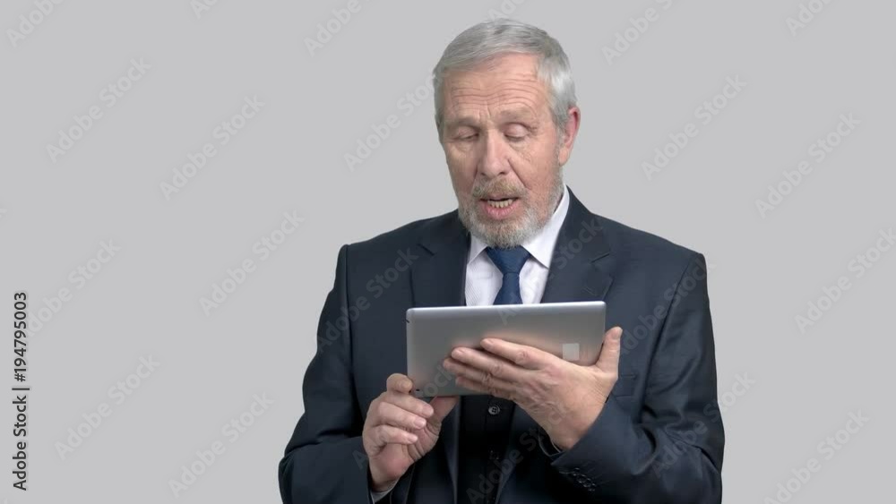 Senior executive talking via internet. Happy grandfather in business suit is talking using computer tablet, grey background. Senior executive talking via internet. Happy grandfather in business suit is talking using computer tablet, grey background.