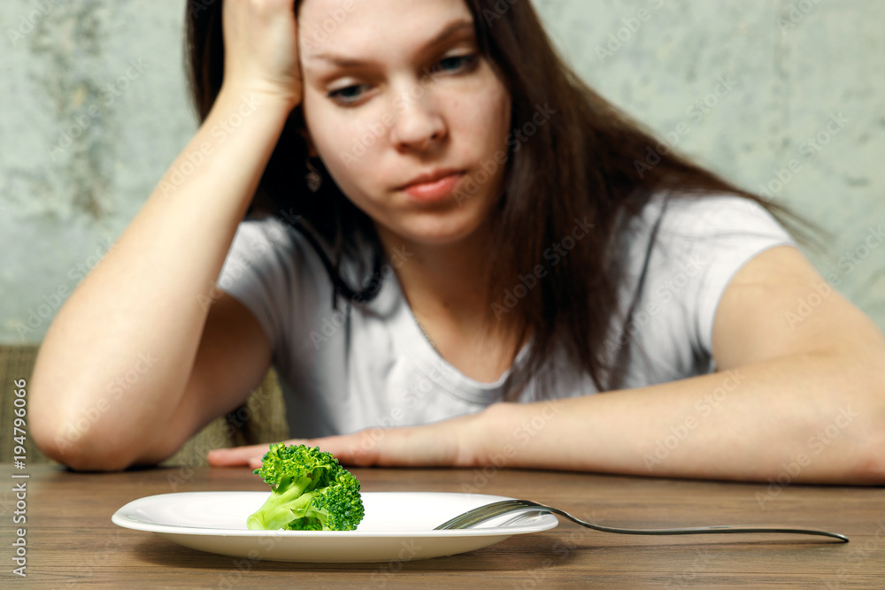 sad young brunette woman dealing with anorexia nervosa or bulimia having small green vegetable on plate. Dieting problems, eating disorder. sad young brunette woman dealing with anorexia nervosa or bulimia having small green vegetable on plate. Dieting problems, eating disorder.