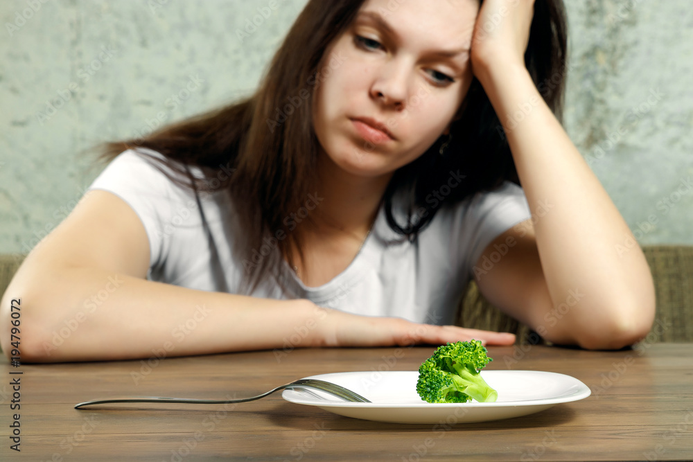 sad young brunette woman dealing with anorexia nervosa or bulimia having small green vegetable on plate. Dieting problems, eating disorder. sad young brunette woman dealing with anorexia nervosa or bulimia having small green vegetable on plate. Dieting problems, eating disorder.