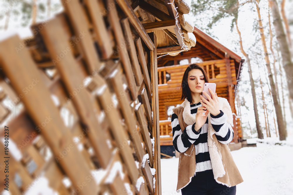 young girl with phone in waistcoat on against of wooden cottage in winter forest