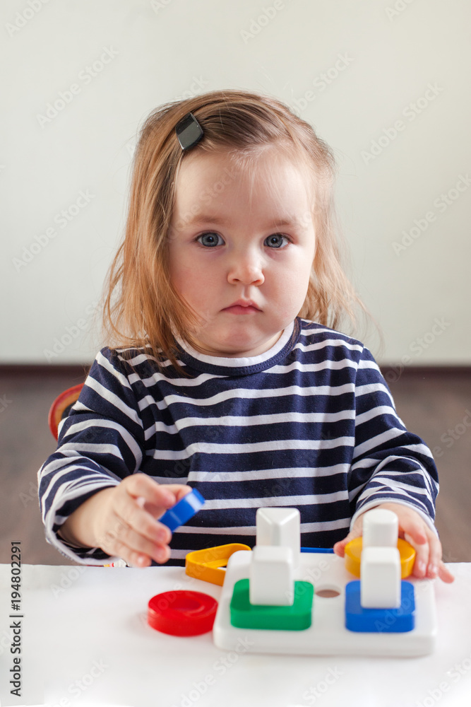 girl 1,5 years old with long hair in a striped dress sits at the table and plays with a developing toy, Montessori technique, portrait, look at the camera girl 1,5 years old with long hair in a striped dress sits at the table and plays with a developing toy, Montessori technique, portrait, look at the camera