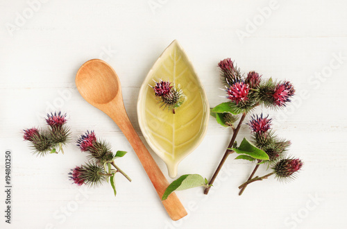 Purple burdock flower heads, plate with herbal oil, top view white wooden table. Natural beauty care treatment. 