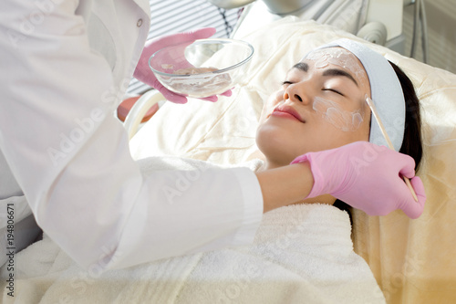 Beautiful young woman wearing headband and bathrobe lying on treatment table while unrecognizable beautician applying moisturizing mask on her face