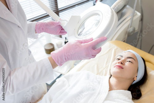 Unrecognizable beautician wearing white coat and rubber gloves using magnifying lamp while examining facial skin of pretty young client lying on treatment table
