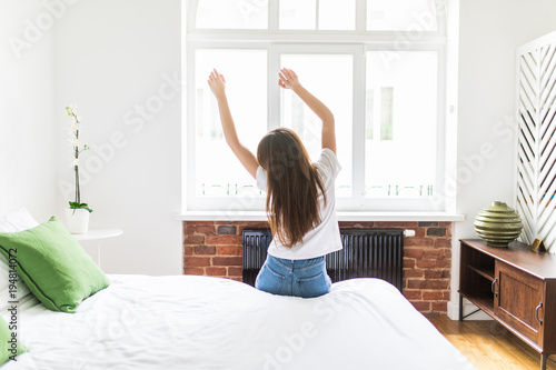 Woman standing in her bed near the window while stretching after waking up with sunrise at morning, back view